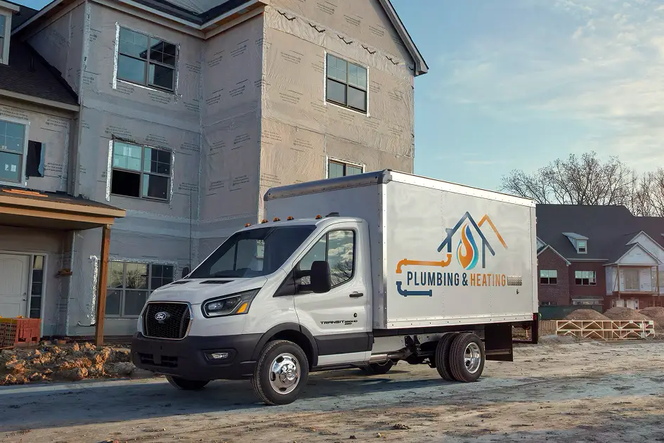 White Ford Transit chassis cab with a dry-freight box body in plumbing contractor livery at a residential construction site